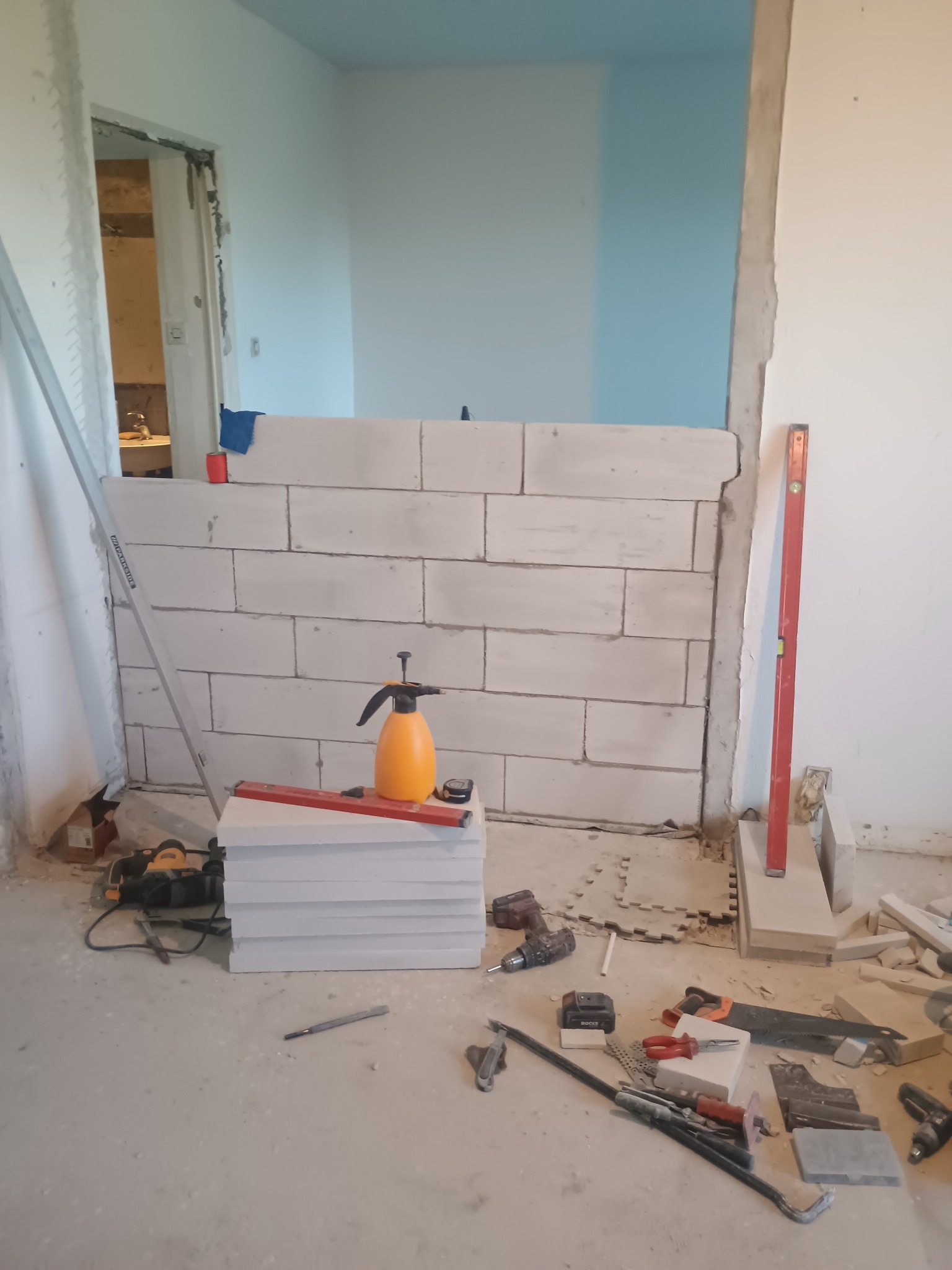 Wznoszenie ściany działowej z bloczków betonu komórkowego. Widoczne narzędzia: poziomica, wiertarka, kielnia. Fragment otwartego przejścia do innego pomieszczenia.