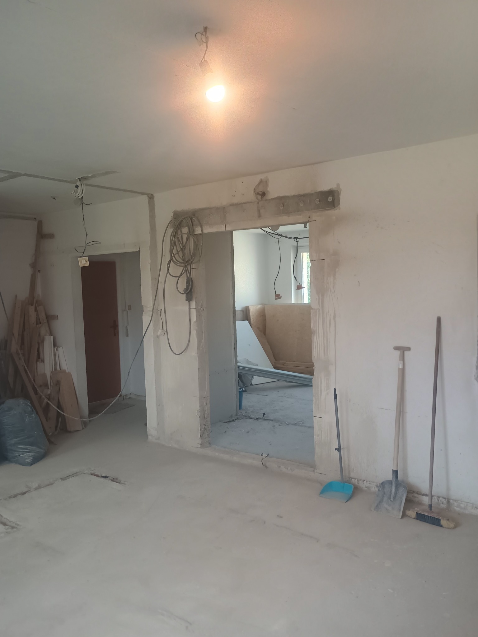 Wnętrze w trakcie remontu: surowy beton, narzędzia (szpadel, zmiotka), widoczne kable i instalacje. W tle przygotowane płyty. Ściany w stanie surowym.