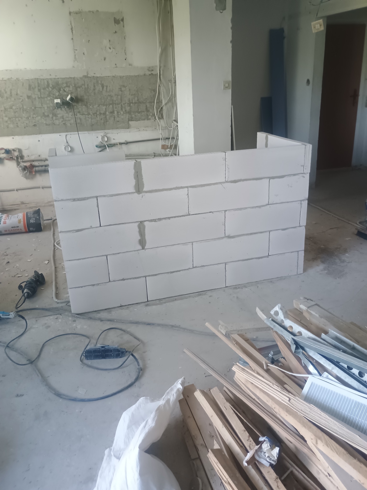 Wnętrze w trakcie remontu: murowana ścianka działowa z białych bloczków, widoczne narzędzia i materiały budowlane, ślady po instalacjach na ścianie.