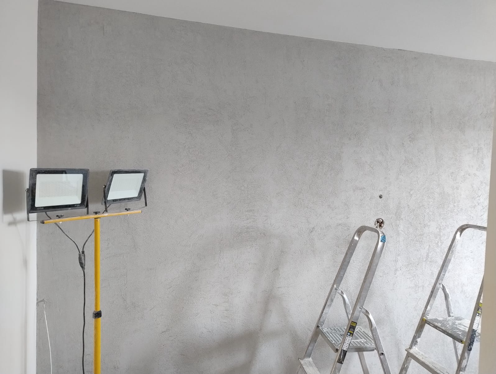 Ściana po malowaniu dekoracyjnym w odcieniach szarości, oświetlona lampami budowlanymi, z drabinami w tle. Efekt wykończenia wnętrza.