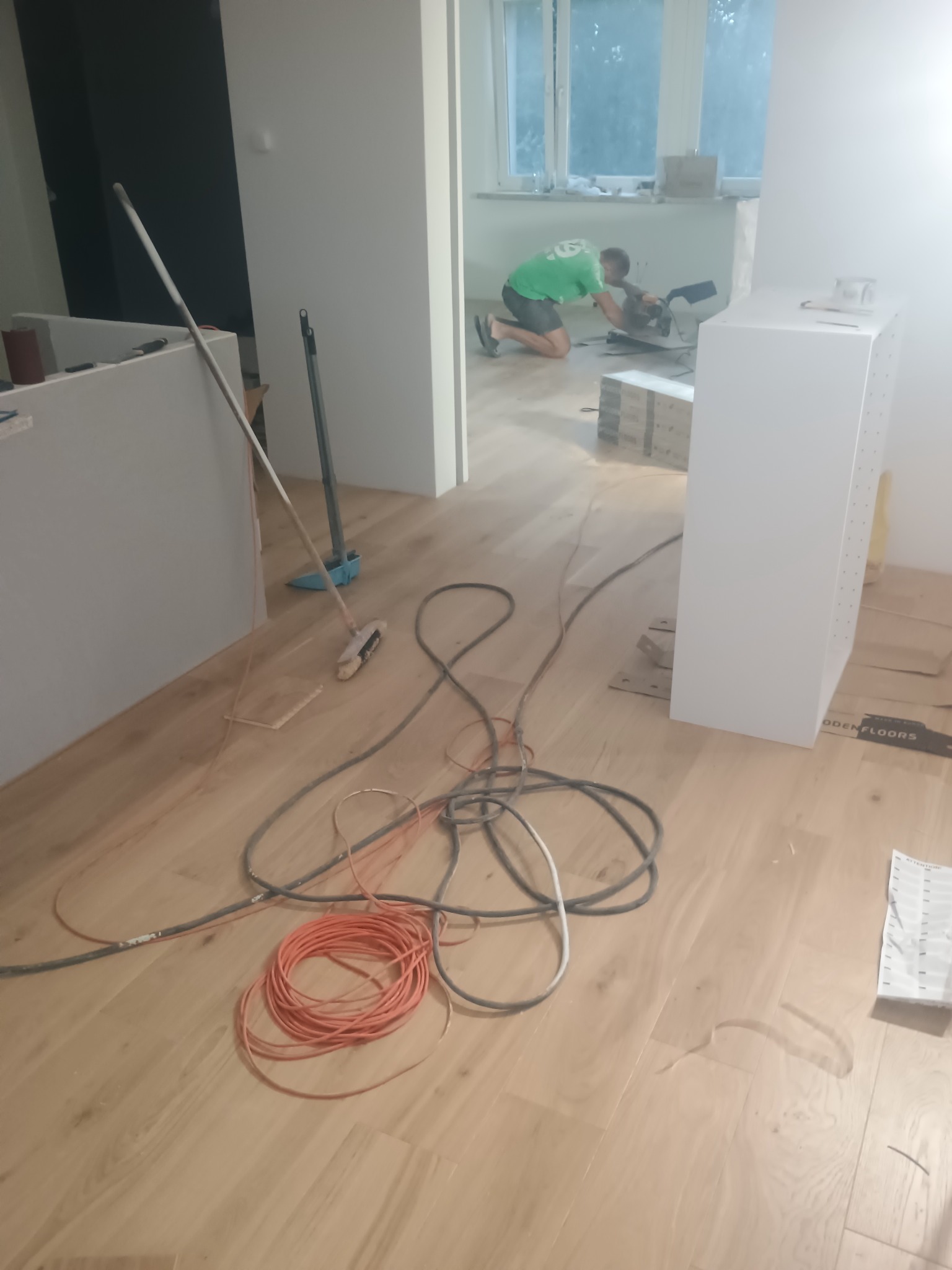 Montaż szafki na drewnianej podłodze, w tle pracownik przycina element. Widoczne kable, narzędzia i akcesoria montażowe. Wnętrze w trakcie wykańczania.