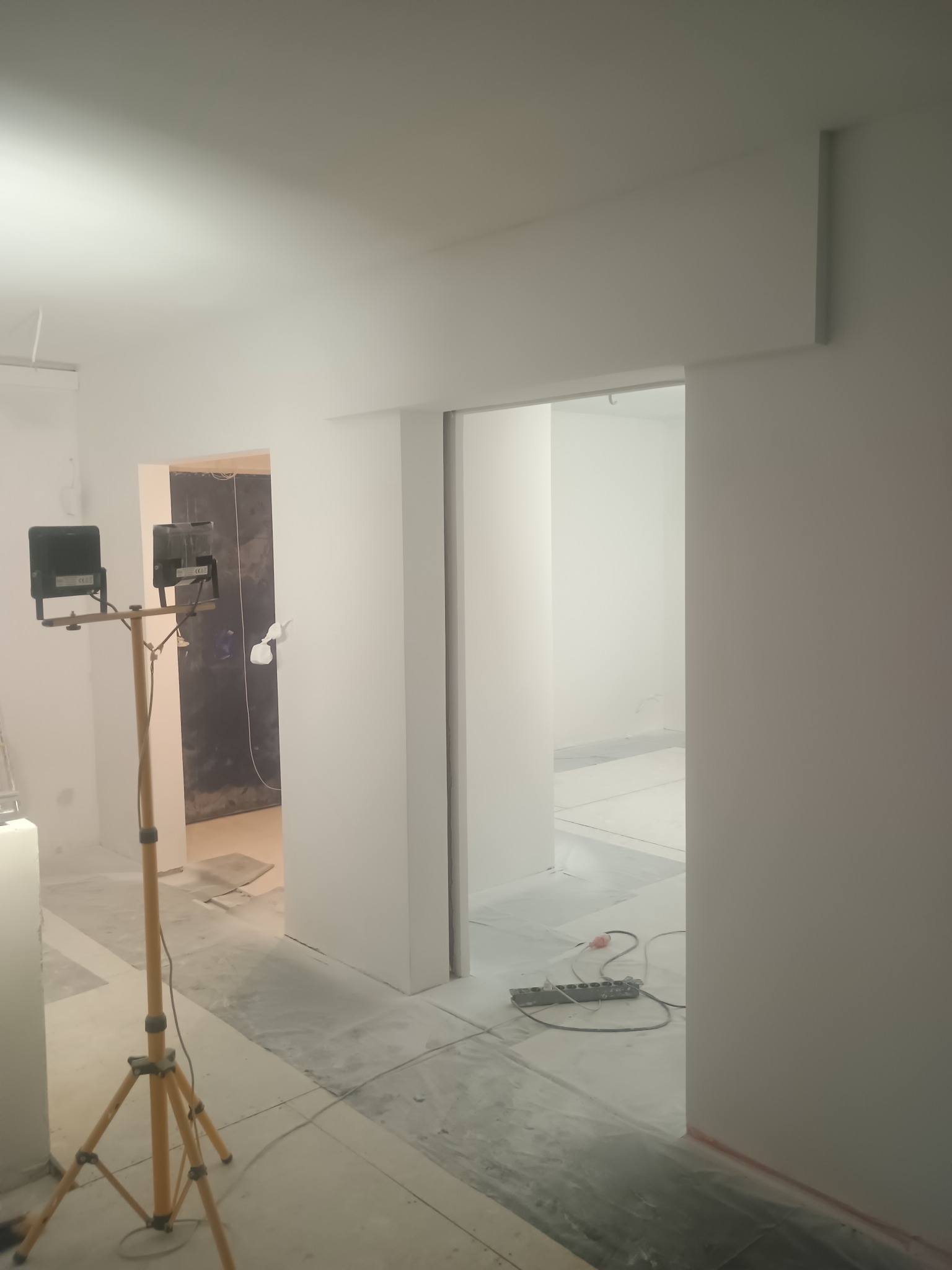 Białe wnętrze w trakcie malowania z lampą na statywie, widoczne zabezpieczenia podłogi folią. W tle otwarte przejście do kolejnego pomieszczenia.