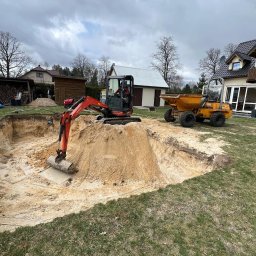 Yukon-Kop - Mini koparka Kubota i wywrotka Thwaites podczas wykopu stawu na posesji z domem. Sprzęt budowlany na trawie, pochmurne niebo.