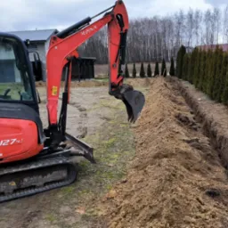 Czerwona mini koparka Kubota U27-4 w trakcie wykonywania wykopu pod drenaż wzdłuż rzędu tuj. Świeżo wykopana ziemia obok rowu. Sprzęt na działce.