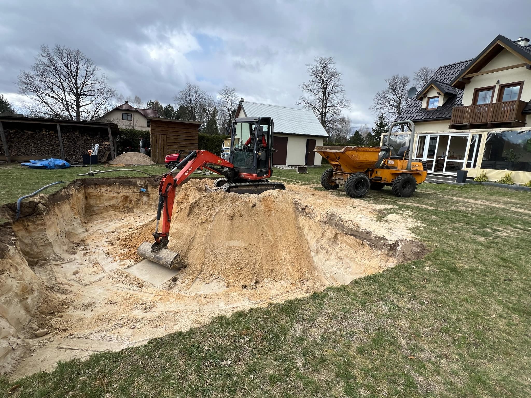 Mini koparka Kubota i wywrotka Thwaites podczas wykopu stawu na posesji z domem. Sprzęt budowlany na trawie, pochmurne niebo.