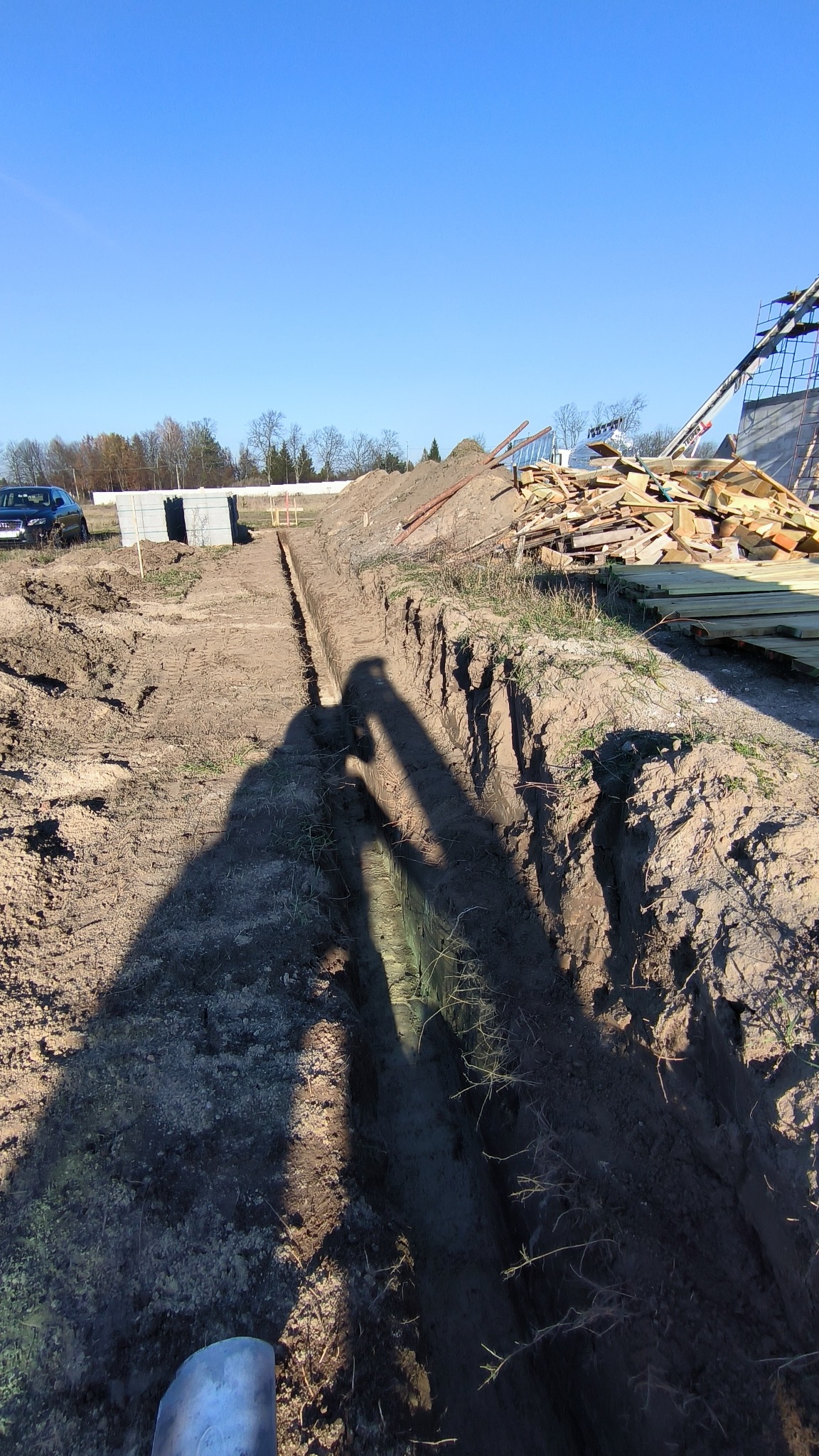 Wykopany wąski rów w ziemi na tle błękitnego nieba, z widocznym cieniem osoby trzymającej szpadel. W tle składowane drewno i częściowo widoczny samochód.