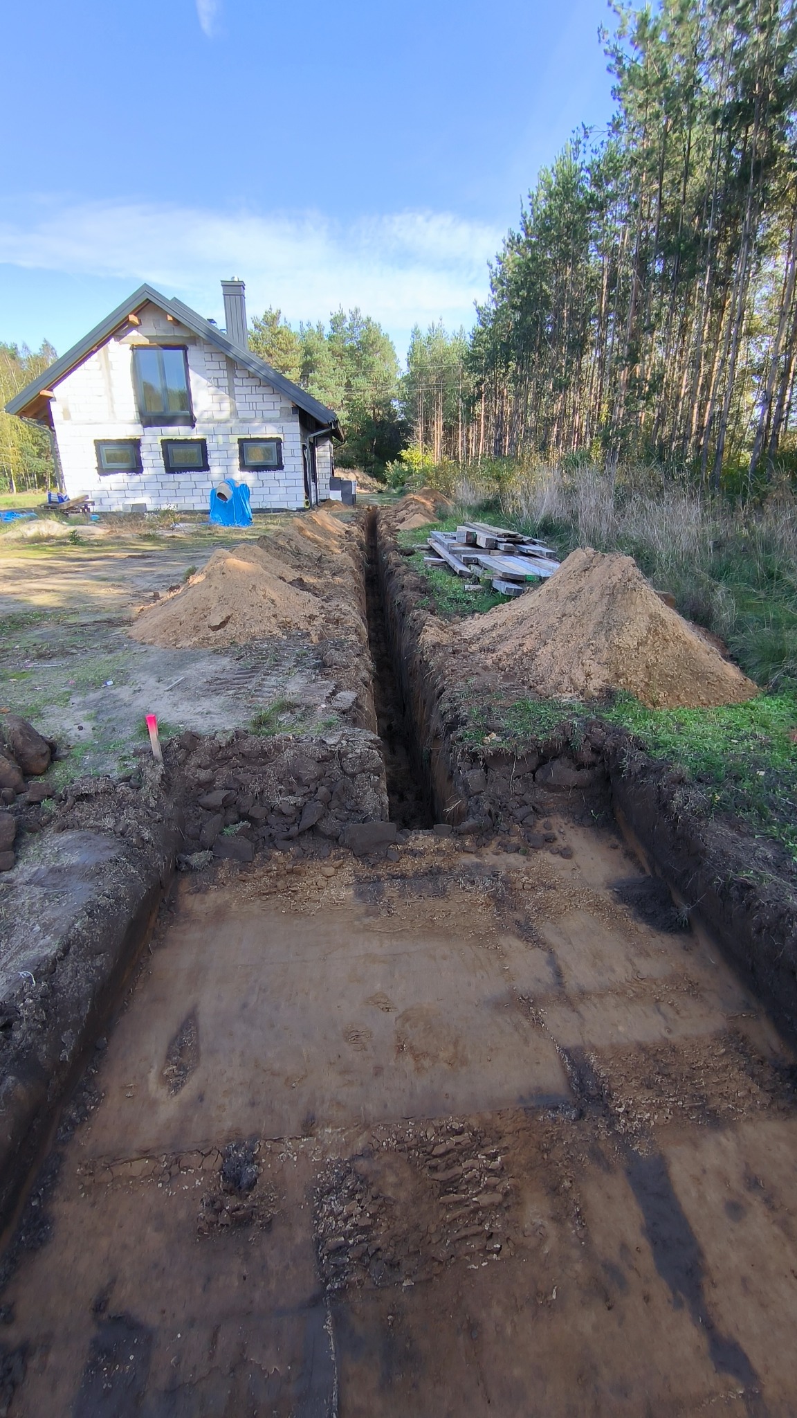 Wykop pod fundamenty domu jednorodzinnego z białego bloczka, widok z góry na wykop i wąski wykop pod instalacje. W tle las iglasty i błękitne niebo.