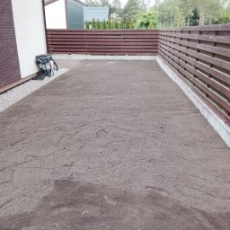 Sztuka ogrodu - Przygotowany teren pod trawnik z rolki, wyrównana ziemia, widoczny drewniany płot i fragment budynku z cegły. Gotowy teren do rozłożenia trawy z rolki.