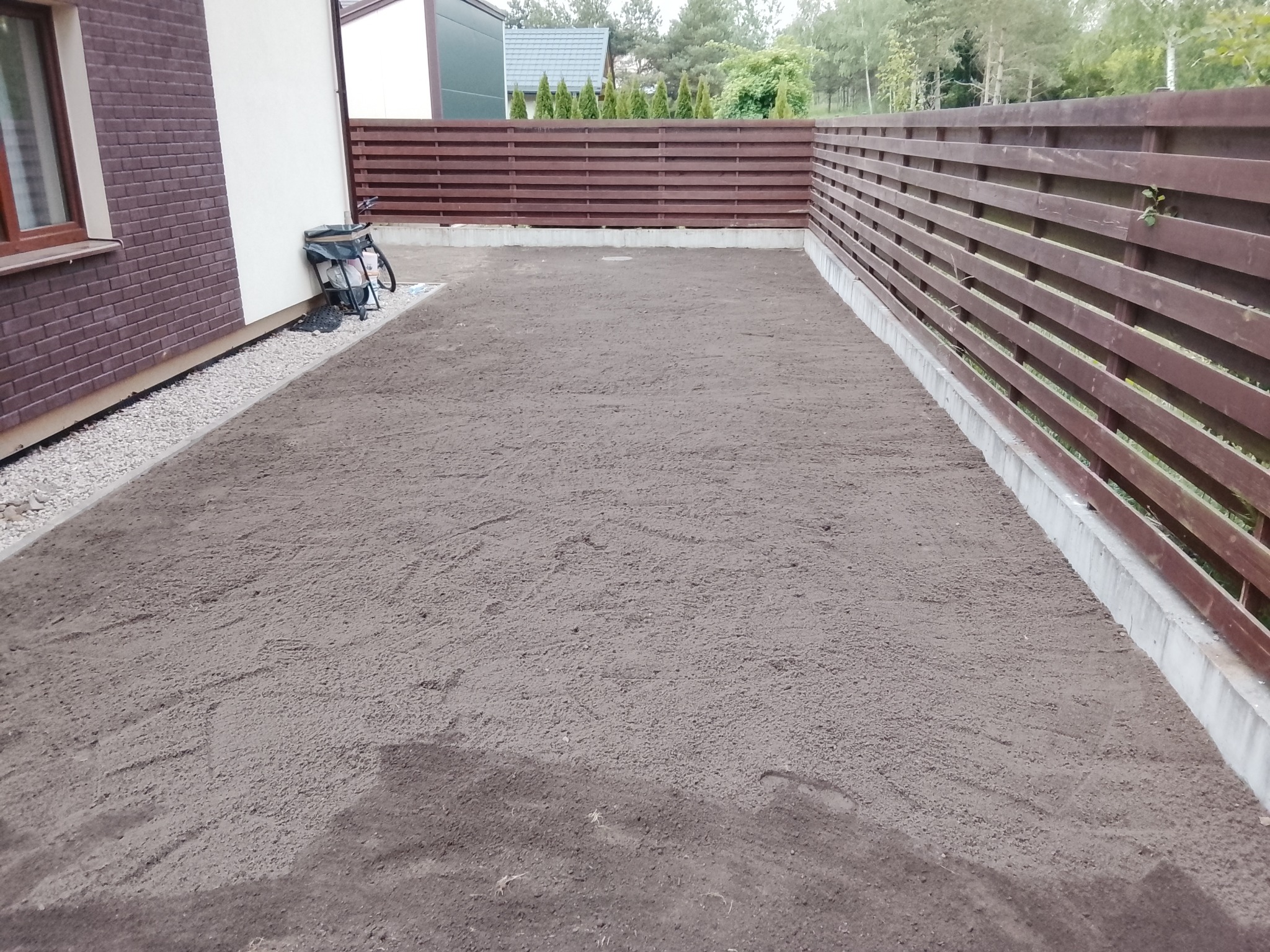 Przygotowany teren pod trawnik z rolki, wyrównana ziemia, widoczny drewniany płot i fragment budynku z cegły. Gotowy teren do rozłożenia trawy z rolki.