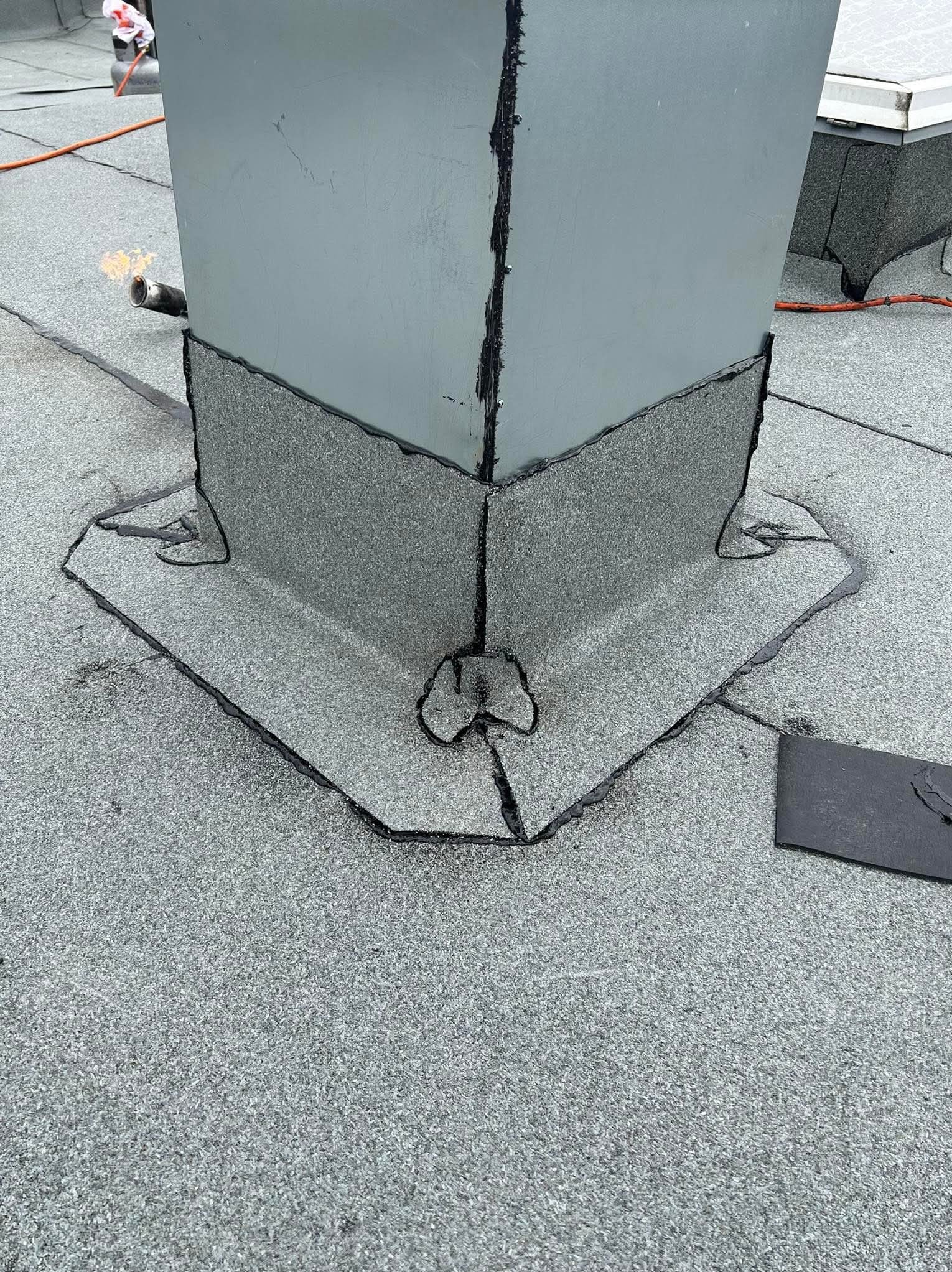 Szczegółowe uszczelnienie komina na płaskim dachu pokrytym papą, z widocznymi spoinami i narzędziem dekarskim w tle.