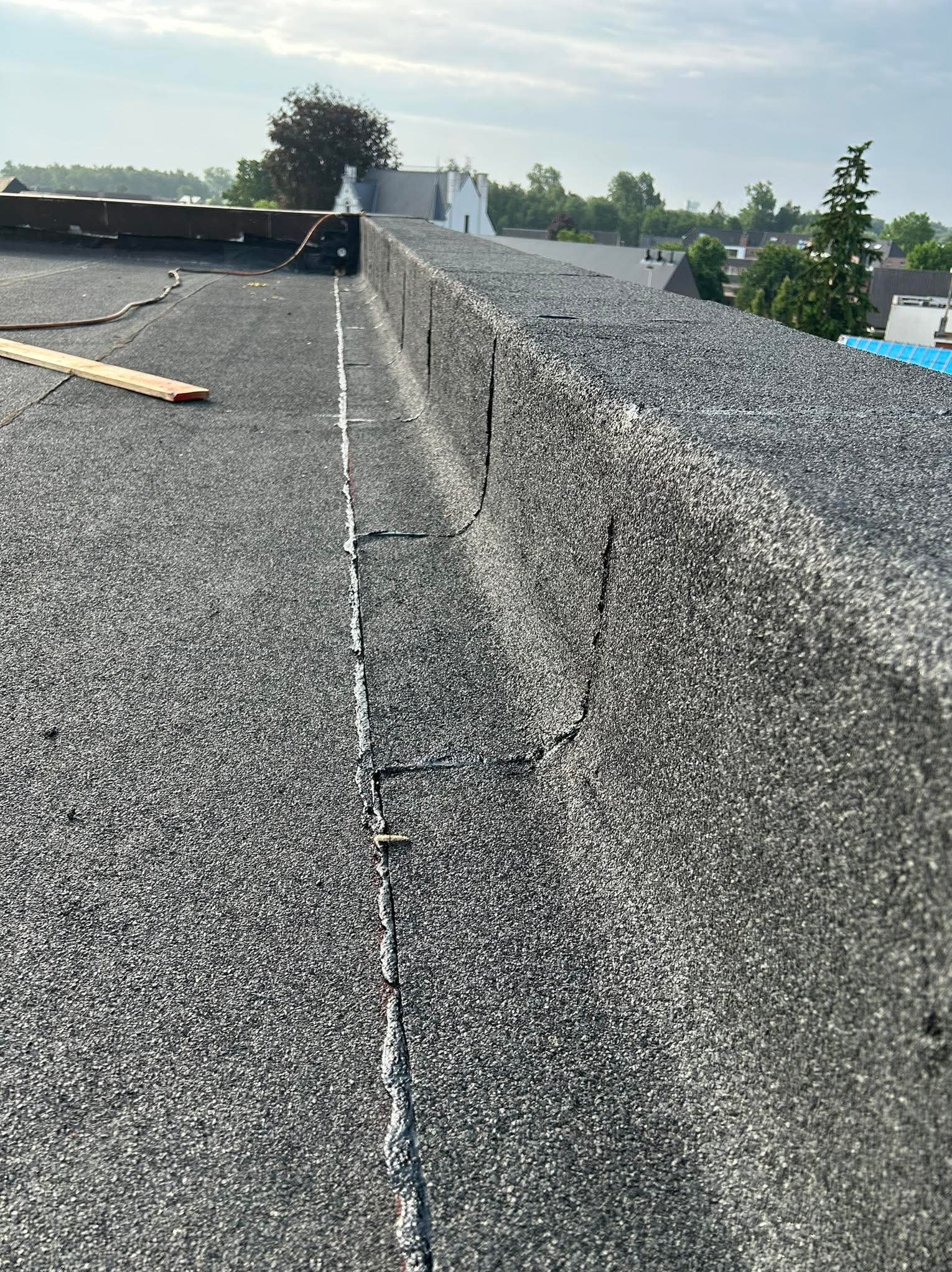Pęknięcia na papie dachowej płaskiego dachu, widoczne uszkodzenia i linia łączenia. W tle zabudowa jednorodzinna i drzewa.