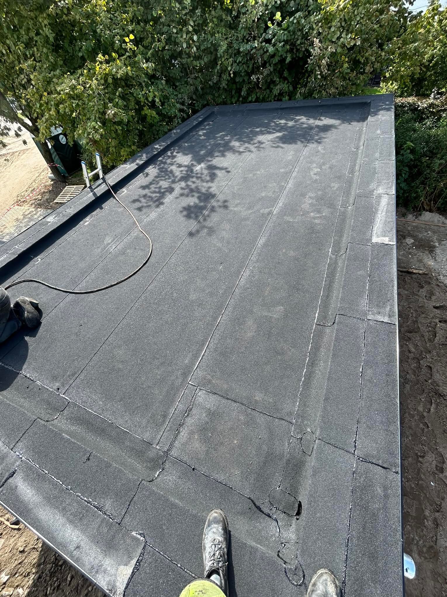 Wykończony dach pokryty papą termozgrzewalną, widoczne łączenia i fragmenty butów dekarza. W tle zieleń drzew i drabina. Ujęcie z góry.