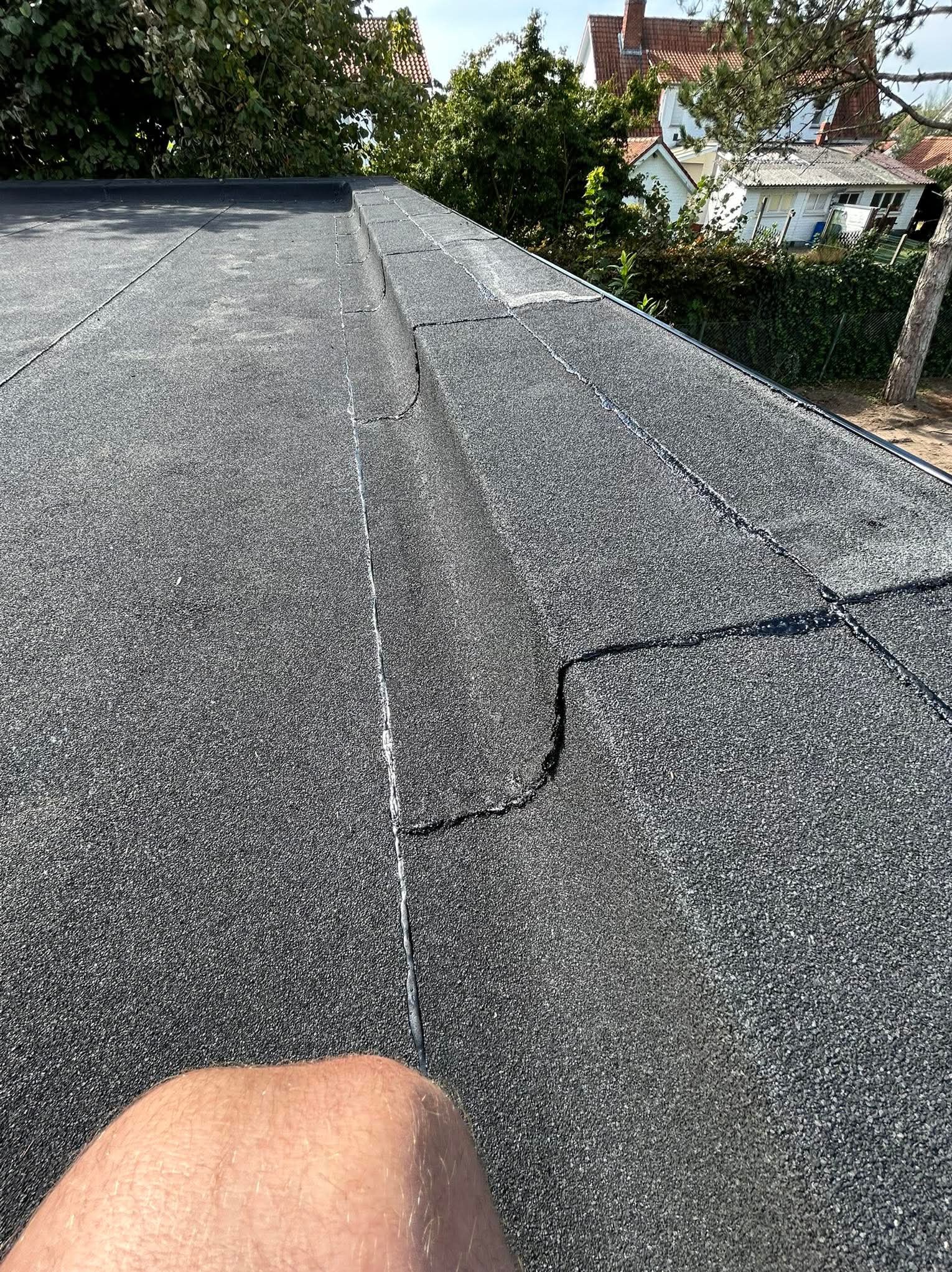 Uszkodzony, płaski dach pokryty papą z widocznymi pęknięciami i naprawami. W tle zieleń drzew i zabudowania mieszkalne.