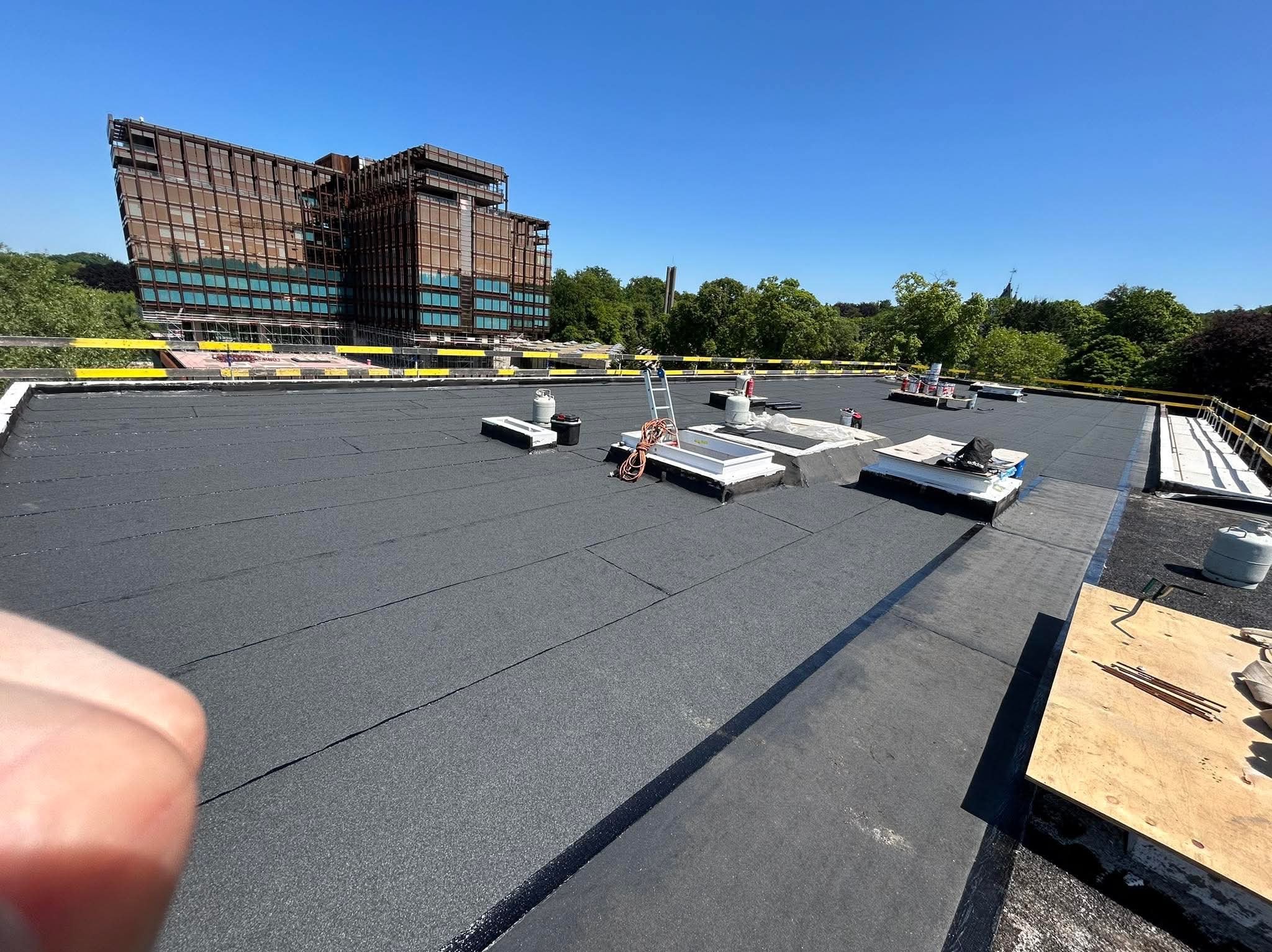 Czarny, płaski dach pokryty papą termozgrzewalną z lukarnami i narzędziami. W tle budynek w stylu industrialnym pod błękitnym niebem.