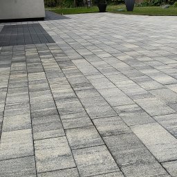 Max - Cleaner Mycie Ciśnieniowe - Ułożona kostka brukowa w odcieniach szarości i beżu, tworząca wzór na podjeździe. Widoczne donice z roślinami i fragment budynku w tle.