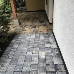 Max - Cleaner Mycie Ciśnieniowe - Ułożona, szara kostka brukowa przed wejściem do budynku z białą elewacją. Część kostki w odcieniach beżu. Widoczny fragment roślinności.