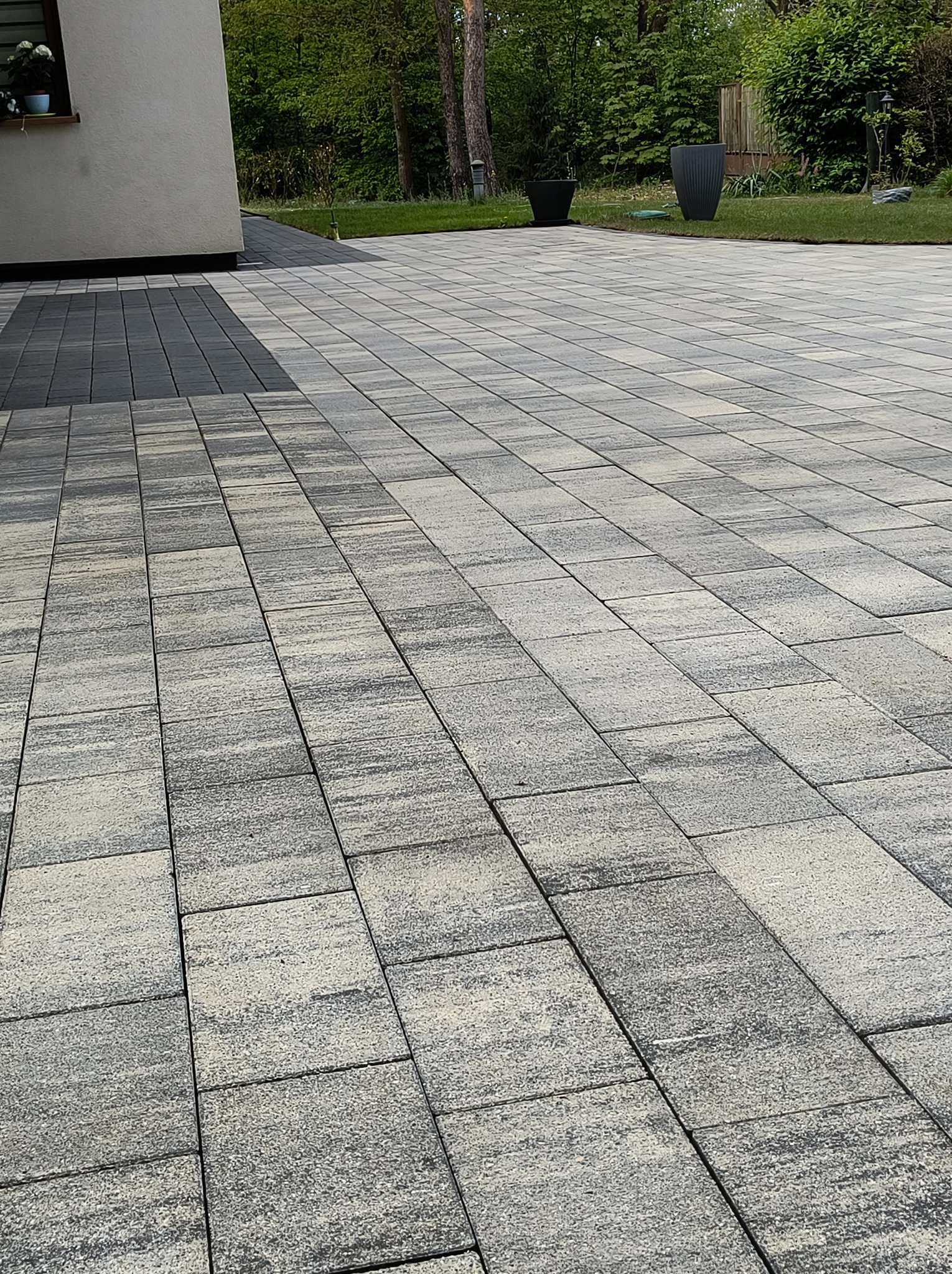 Ułożona kostka brukowa w odcieniach szarości i beżu, tworząca wzór na podjeździe. Widoczne donice z roślinami i fragment budynku w tle.