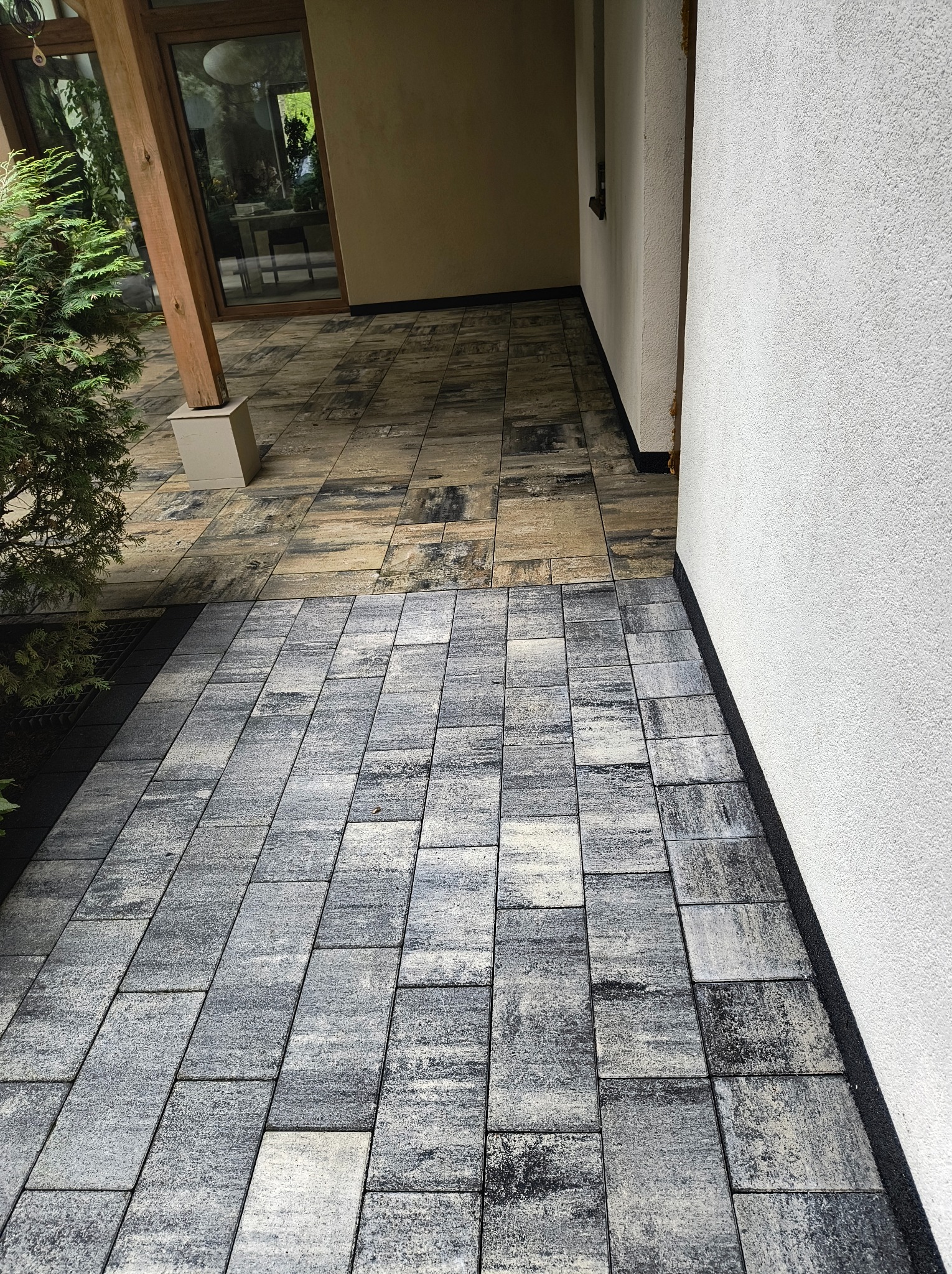 Ułożona, szara kostka brukowa przed wejściem do budynku z białą elewacją. Część kostki w odcieniach beżu. Widoczny fragment roślinności.