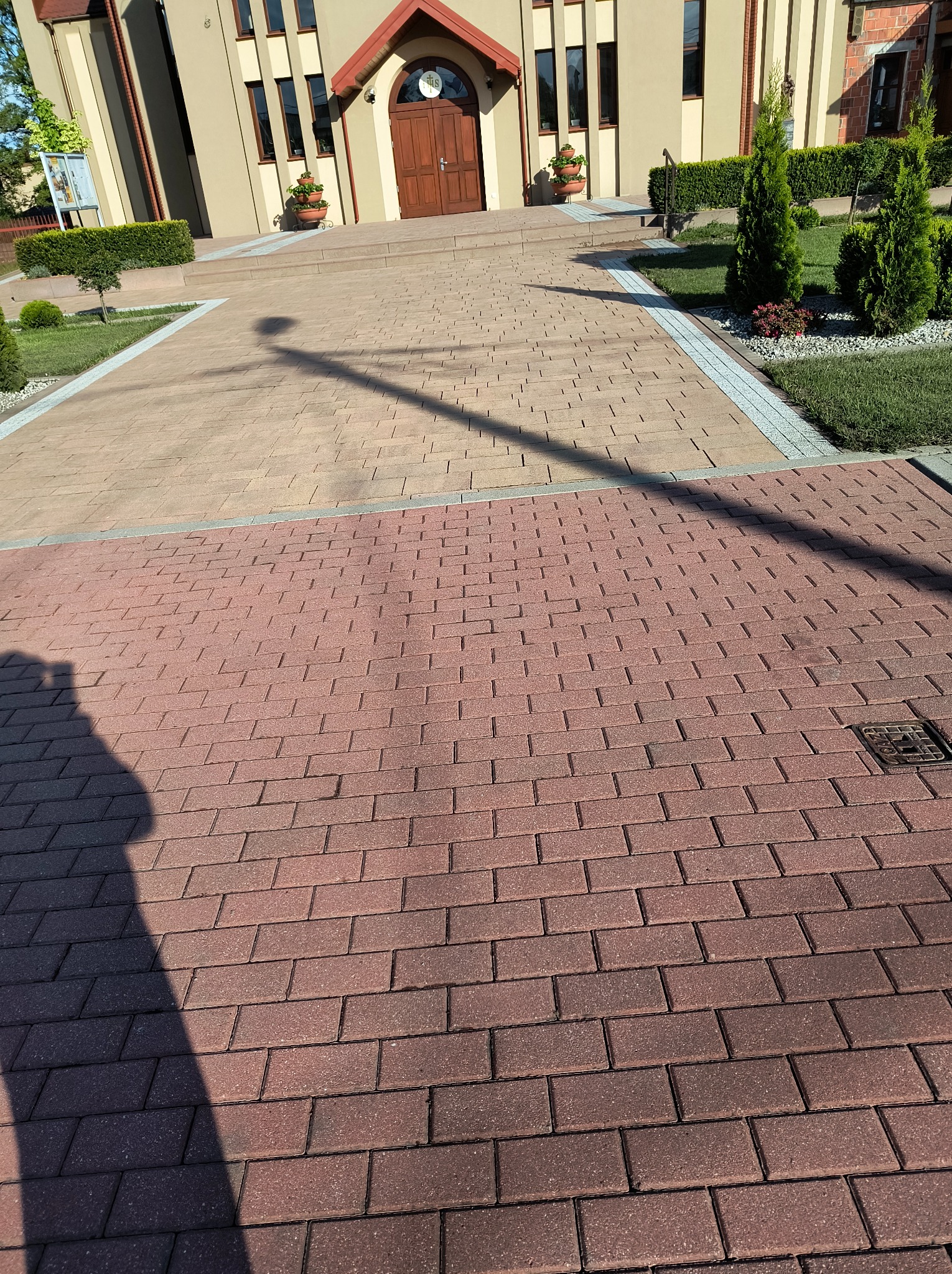 Czerwona kostka brukowa prowadzi do budynku z beżową elewacją. Widoczny cień osoby. Ułożona kostka brukowa w dwóch odcieniach, zieleń wokół.