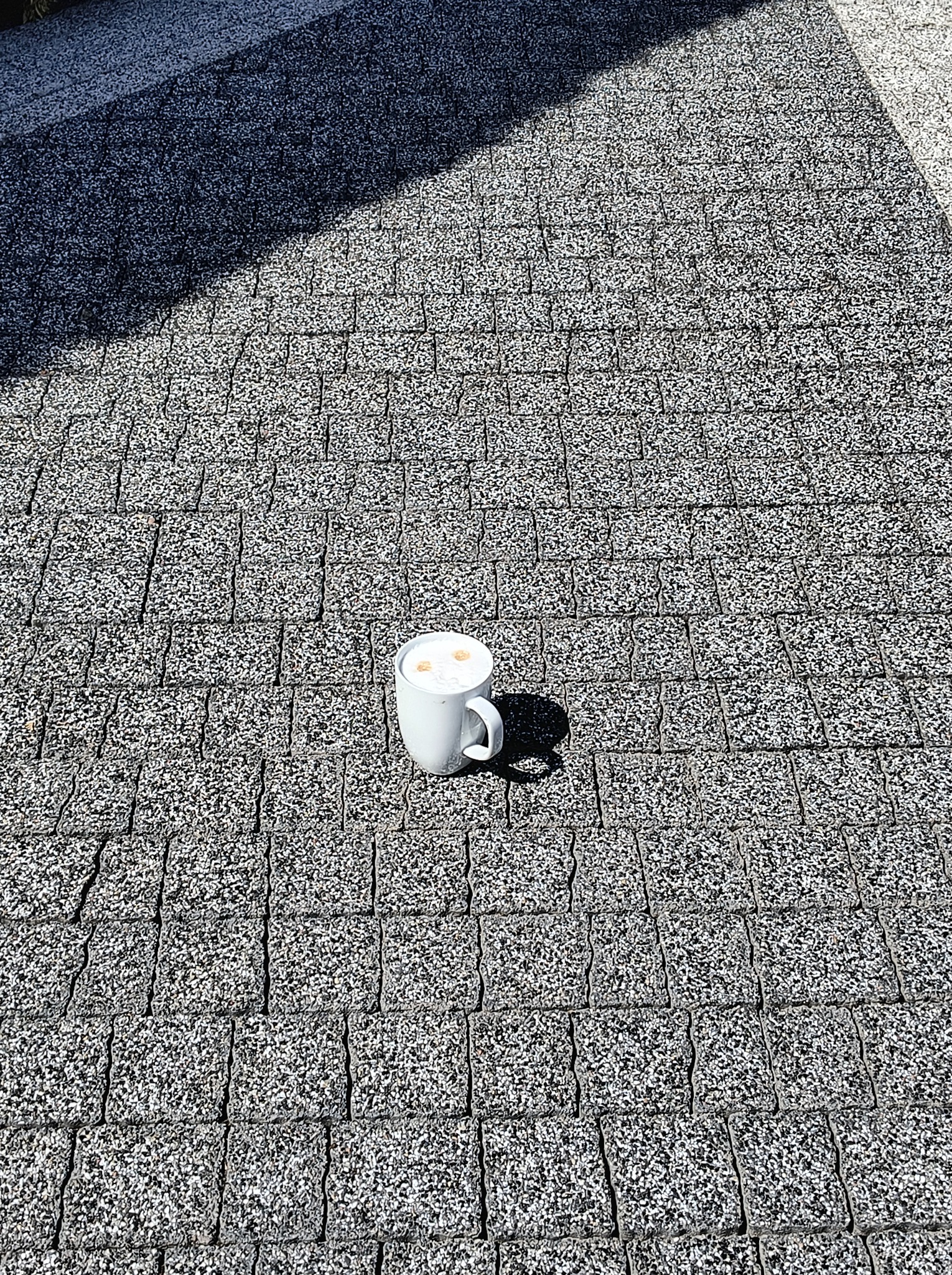 Szara kostka brukowa w słoneczny dzień z białą filiżanką kawy pośrodku, rzucająca cień. Widok z góry na regularny wzór kostki.
