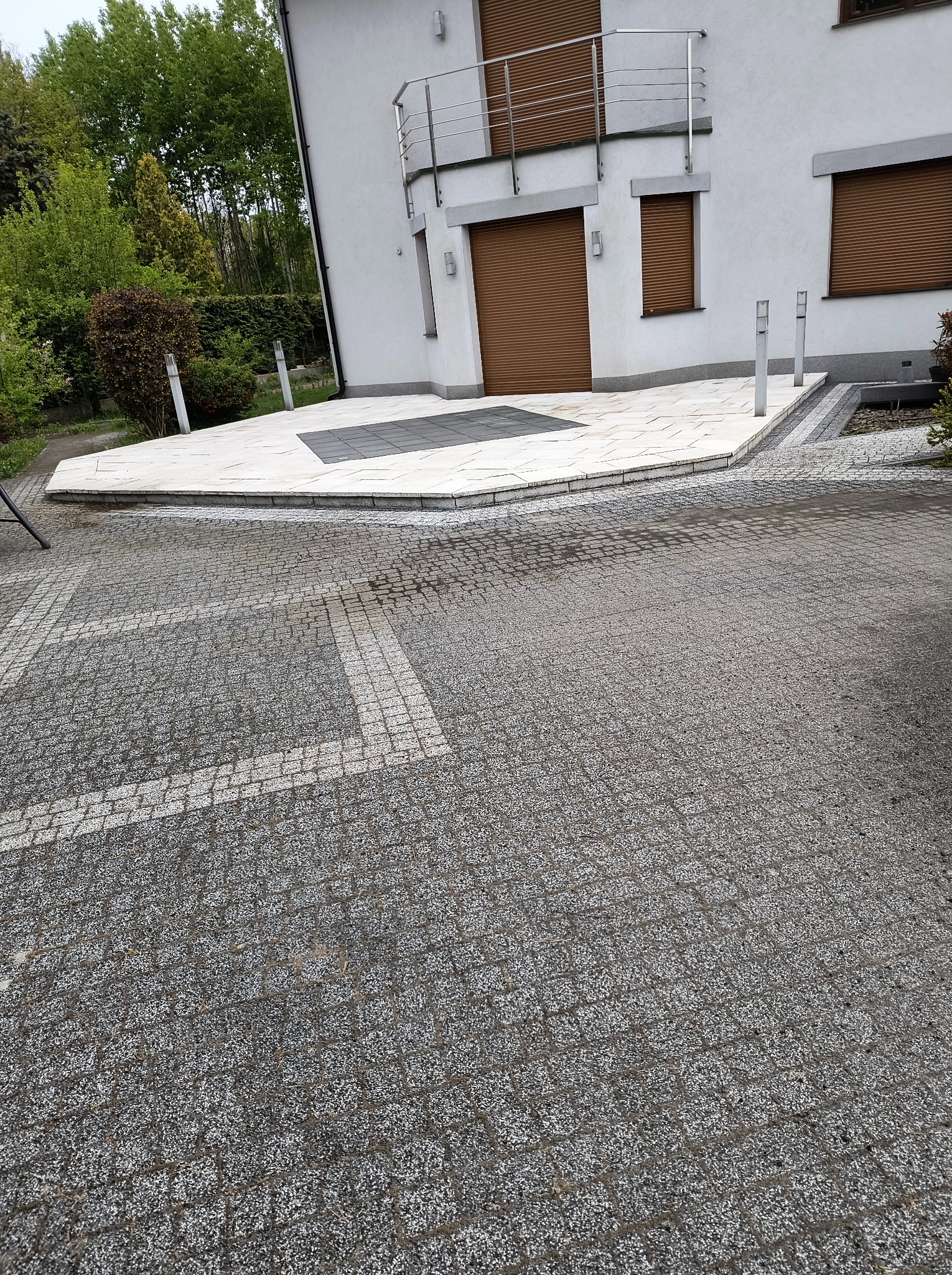 Szara kostka brukowa z białymi pasami tworząca wzór parkingu, obok jasna kostka na tarasie domu z brązowymi roletami w oknach.