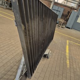 Krzysztof Wijata - Surowa, metalowa konstrukcja przypominająca fragment ogrodzenia panelowego w warsztacie, widoczna z boku, z żółtymi liniami na podłodze z kostki brukowej.