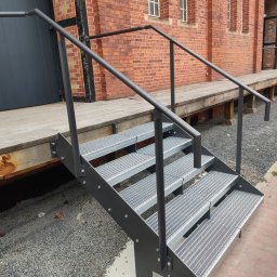 Krzysztof Wijata - Zewnętrzne, metalowe schody kratowe z czarną balustradą, prowadzące do drewnianego podestu przy ceglanym budynku. Widoczne detale mocowań i szara nawierzchnia.