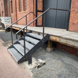 Krzysztof Wijata - Metalowe schody z poręczą prowadzące do drewnianego podestu przy ceglanym budynku. Widoczny gruz i chodnik z kostki.