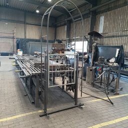 Krzysztof Wijata - Metalowa konstrukcja łukowa w warsztacie, z elementami ogrodzenia na stole. Widoczne narzędzia i materiały, surowe wnętrze hali produkcyjnej. Oświetlenie dzienne.