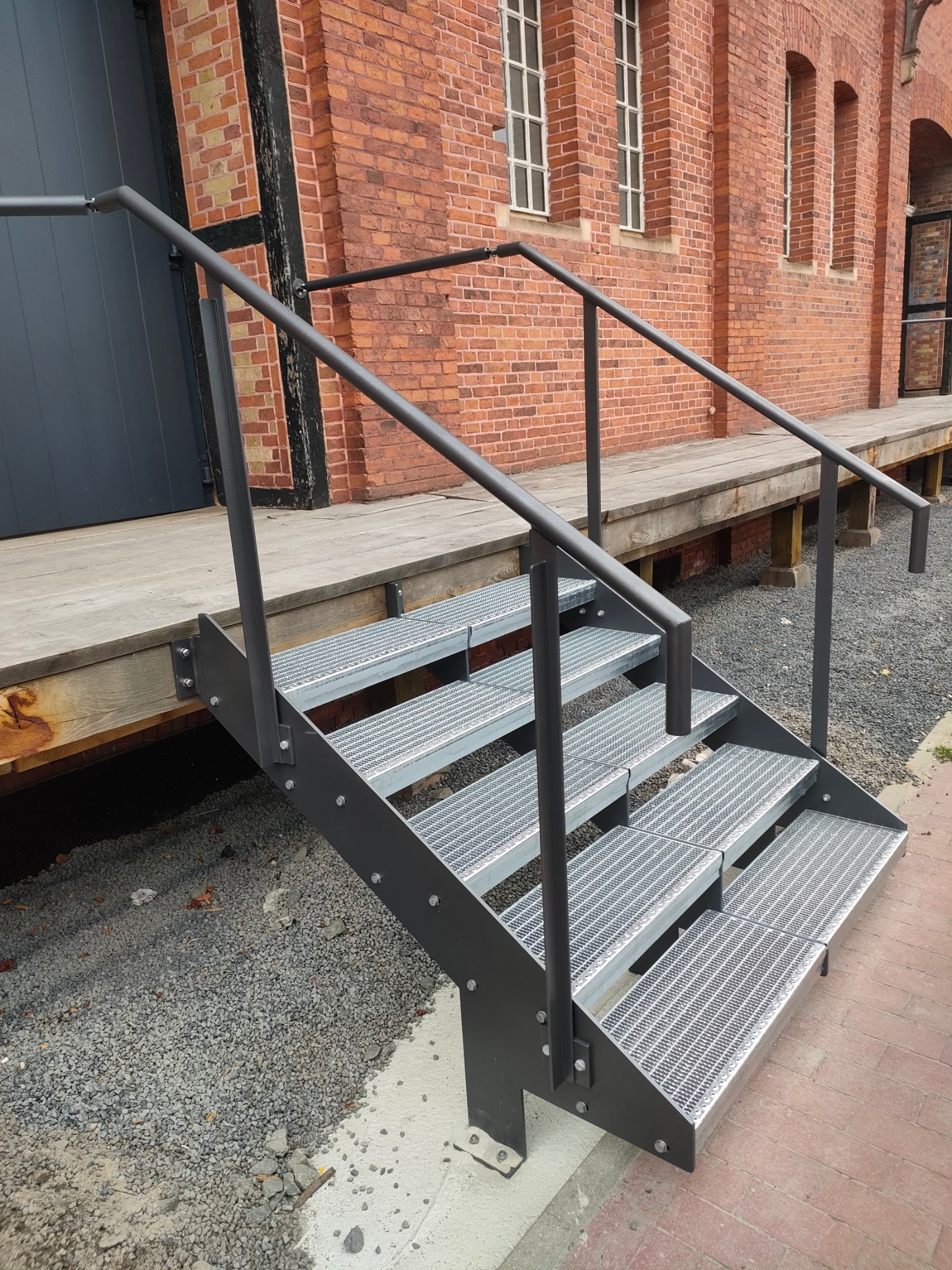 Zewnętrzne, metalowe schody kratowe z czarną balustradą, prowadzące do drewnianego podestu przy ceglanym budynku. Widoczne detale mocowań i szara nawierzchnia.