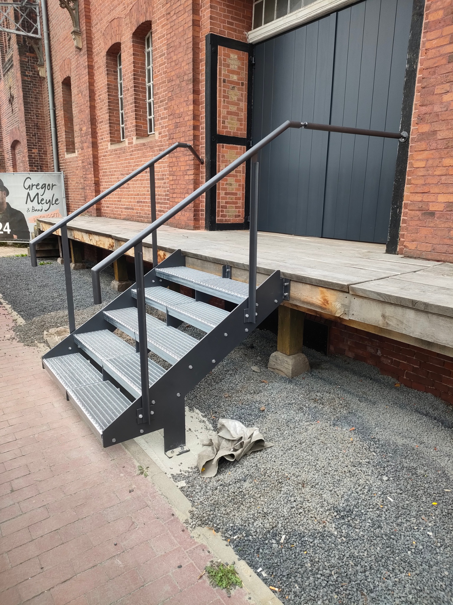 Metalowe schody z poręczą prowadzące do drewnianego podestu przy ceglanym budynku. Widoczny gruz i chodnik z kostki.