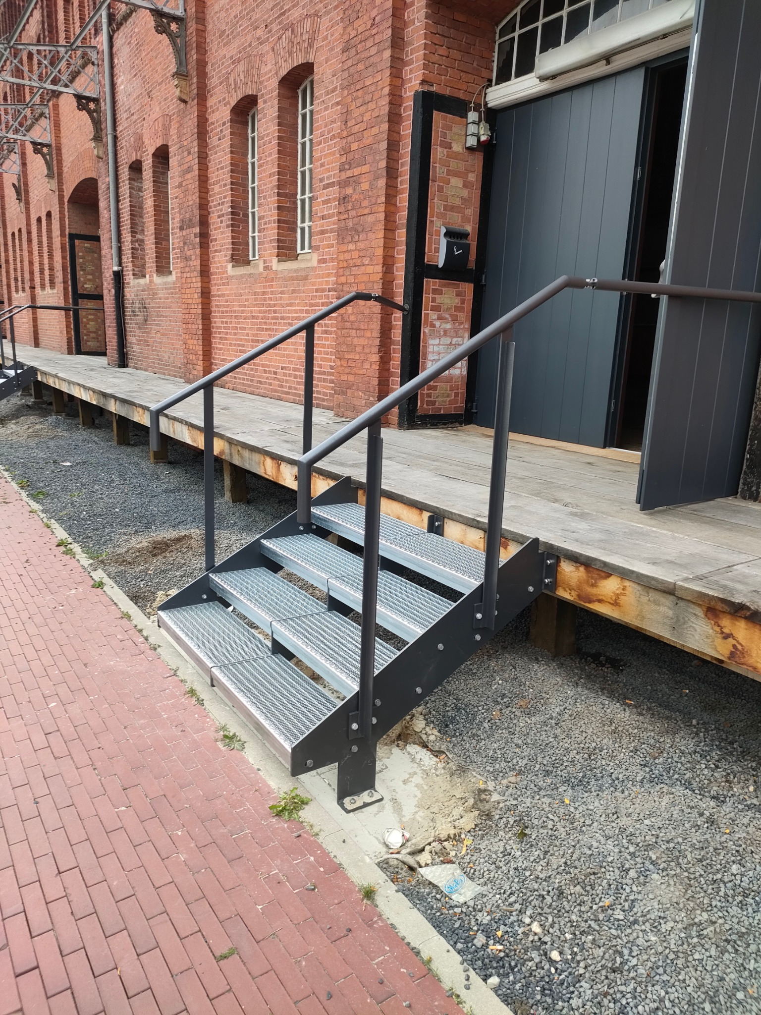 Metalowe schody z poręczą prowadzą do drewnianego podestu przy ceglanym budynku z szarymi, przesuwnymi drzwiami. Widoczne kostki brukowe i żwirowe podłoże.