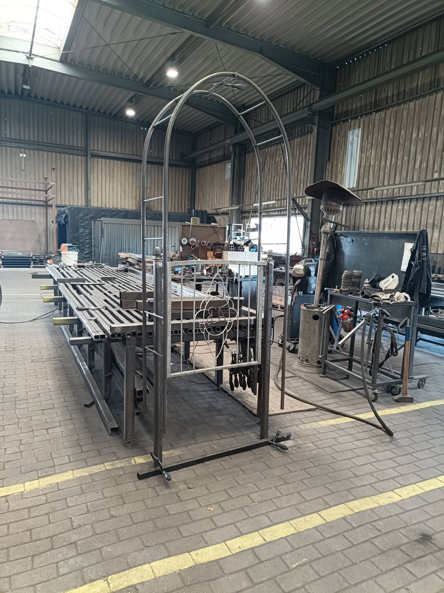 Metalowa konstrukcja łukowa w warsztacie, z elementami ogrodzenia na stole. Widoczne narzędzia i materiały, surowe wnętrze hali produkcyjnej. Oświetlenie dzienne.