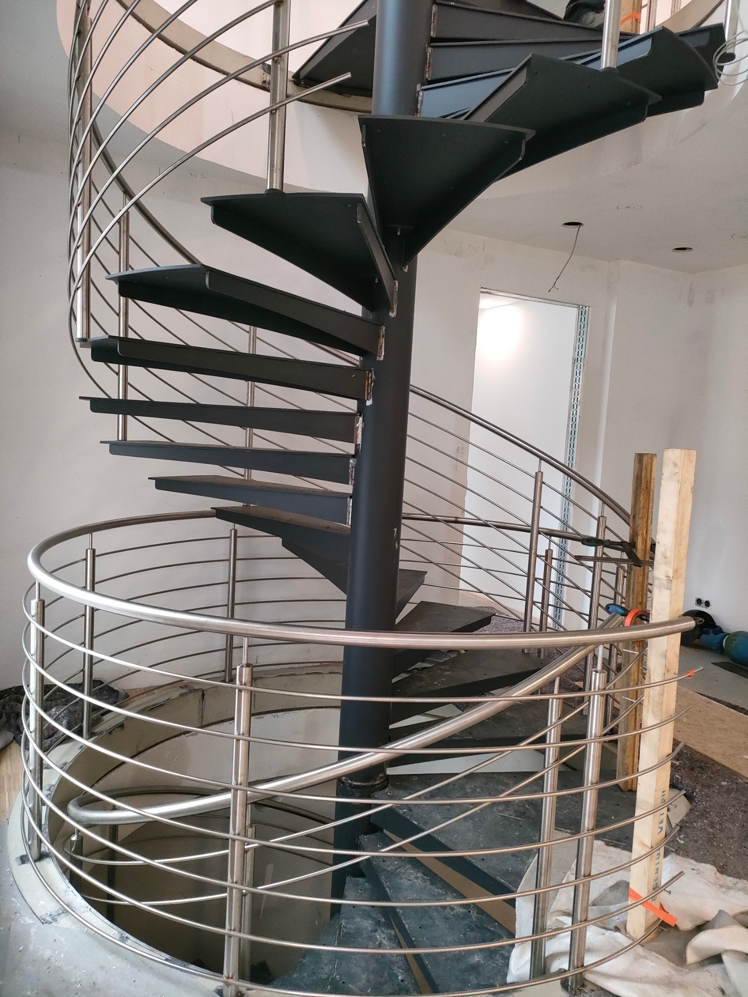Metalowe schody spiralne z czarnymi stopniami i srebrną balustradą, widok z dołu, w surowym wnętrzu. Widoczne niedokończone prace wykończeniowe.