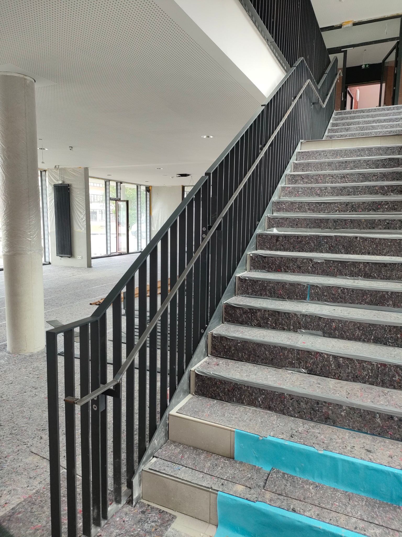 Nowoczesna, czarna balustrada schodowa z metalu, widoczna z dołu, z zabezpieczonymi schodami w budynku w trakcie wykończenia. Widoczne okna i kolumna.