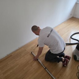 Belago - Pracownik w trakcie czyszczenia parkietu przy ścianie odkurzaczem przemysłowym. Widoczny fragment podłogi i ściana z listwami przypodłogowymi.