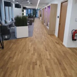 Jasna, drewniana podłoga ułożona w jodełkę w nowoczesnym biurze z elementami zieleni i strefą relaksu. Widoczny fragment gaśnicy na ścianie.