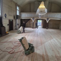 Belago - Cyklinowanie drewnianej podłogi w kościele w Zduńskiej Woli. Parkieciarz w masce podczas pracy. Widoczny proszek i maszyny do cyklinowania. Jasne drewno.