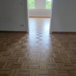 Belago - Wzór parkietu w jodełkę na podłodze w pustym pokoju, widok z perspektywy wejścia. Jasne ściany i okno w tle. Parkiet ułożony w kwadraty z różnych odcieni drewna.