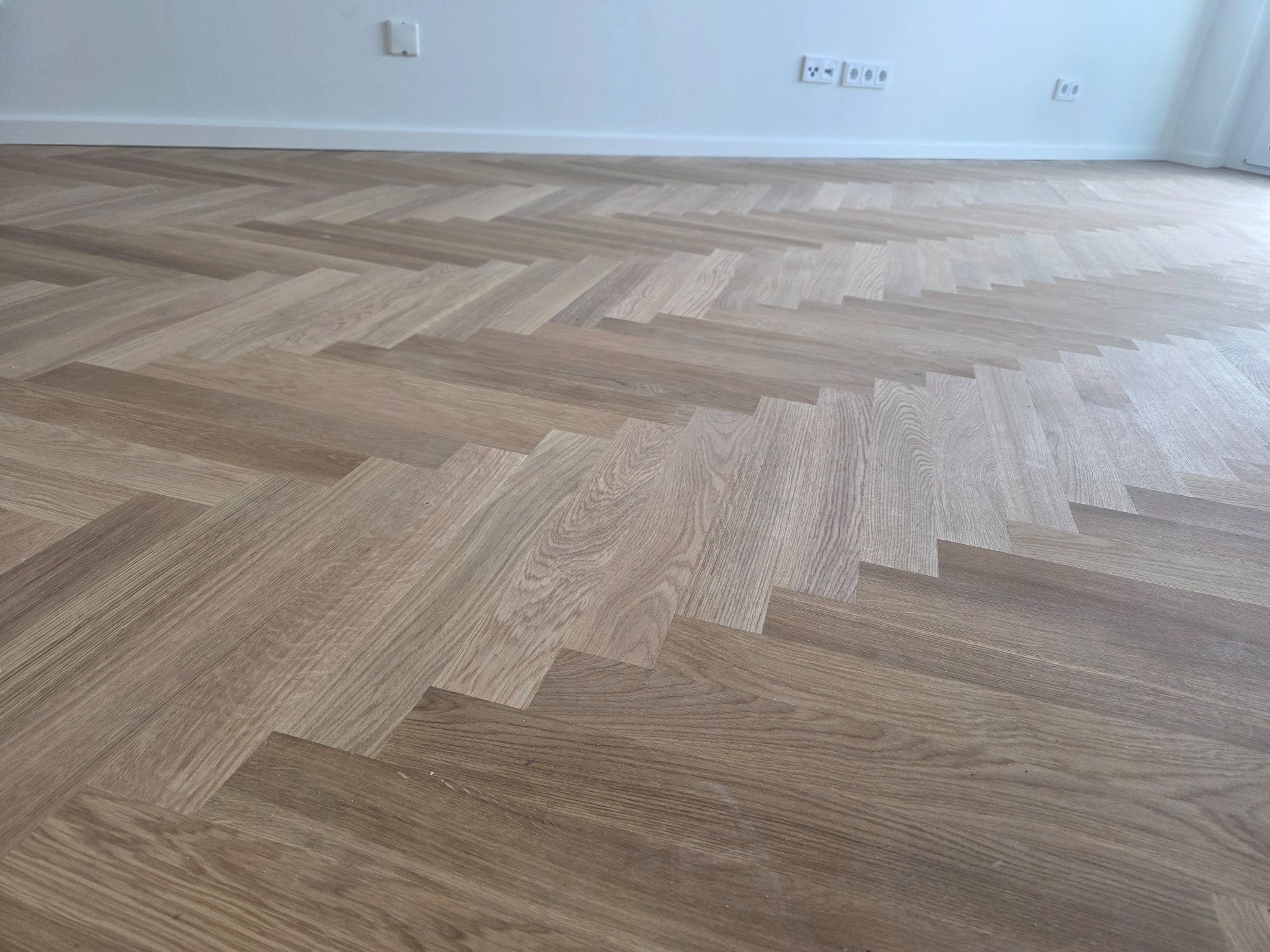 Ułożona podłoga z drewnianego parkietu w jodełkę, z widoczną strukturą drewna, w jasnym pomieszczeniu z białymi ścianami i gniazdkami.