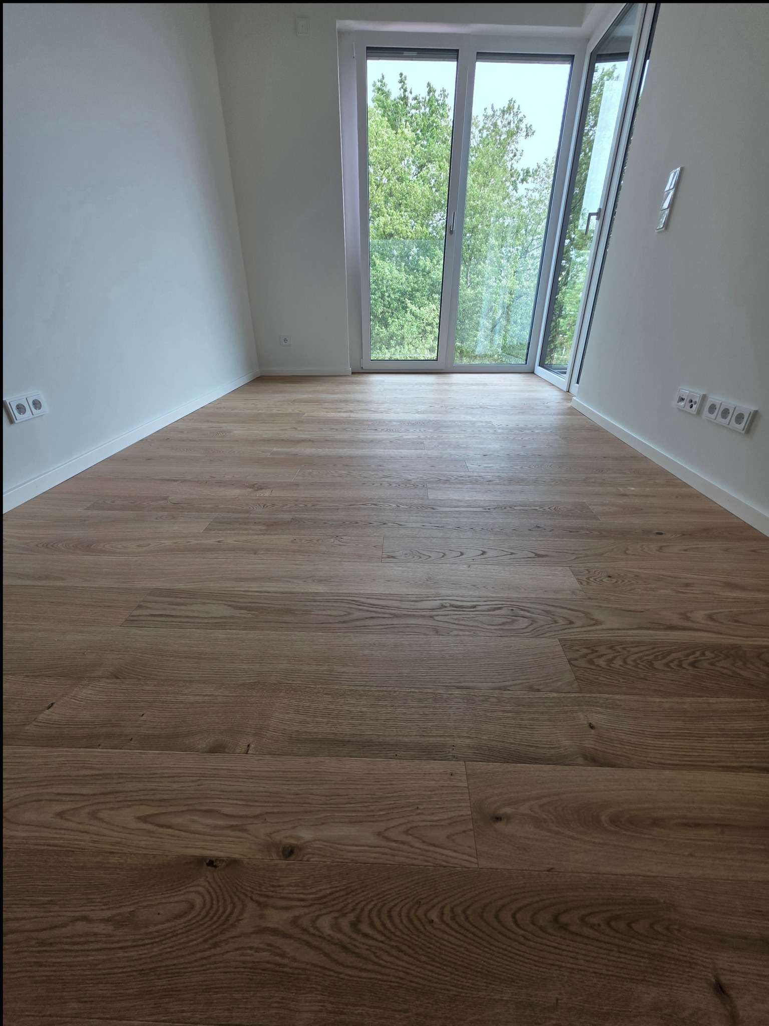 Jasna, drewniana podłoga ułożona w pustym pokoju z dużym oknem balkonowym. Widoczne listwy przypodłogowe i gniazdka elektryczne na białych ścianach.