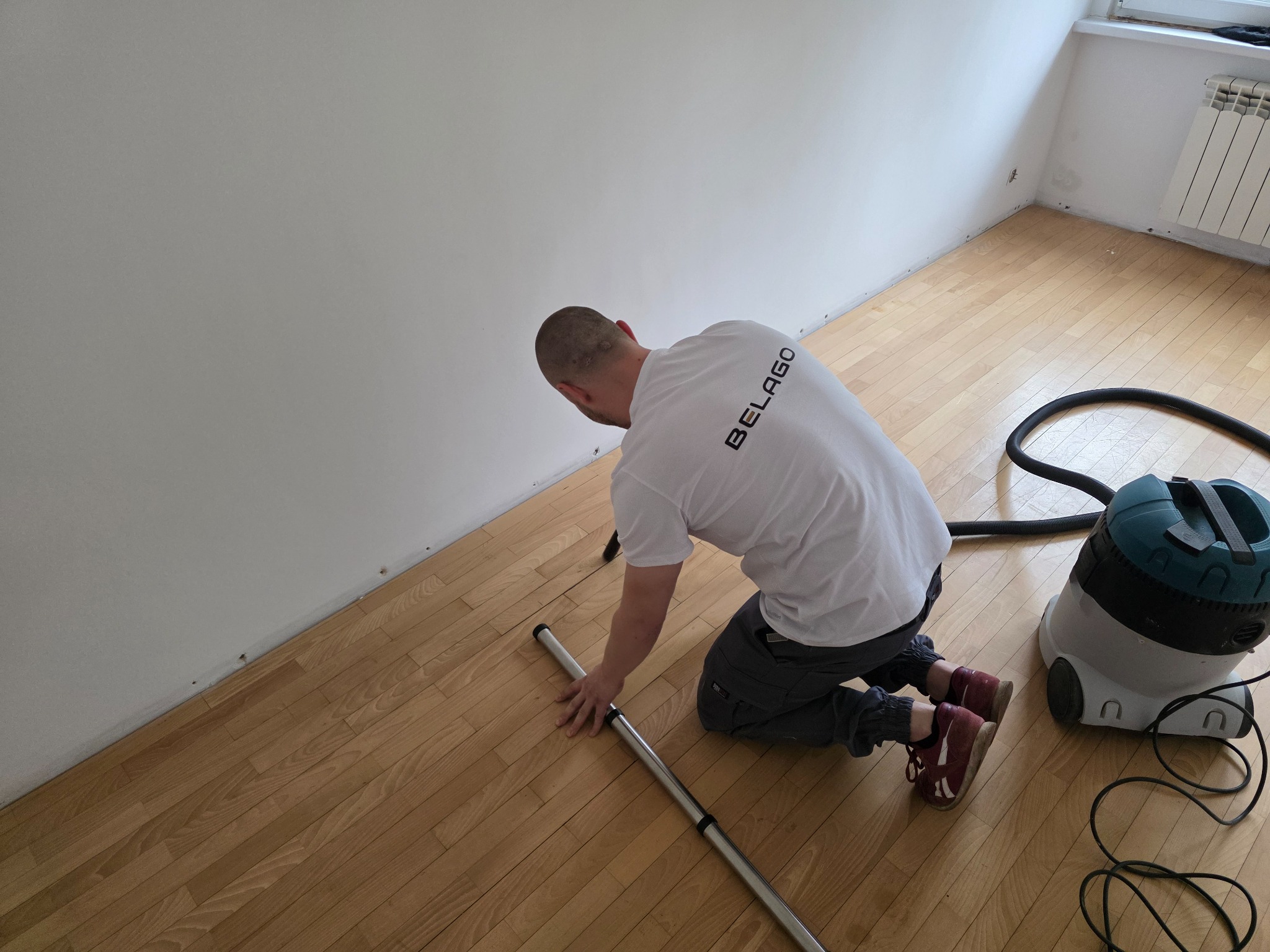 Pracownik w trakcie czyszczenia parkietu przy ścianie odkurzaczem przemysłowym. Widoczny fragment podłogi i ściana z listwami przypodłogowymi.