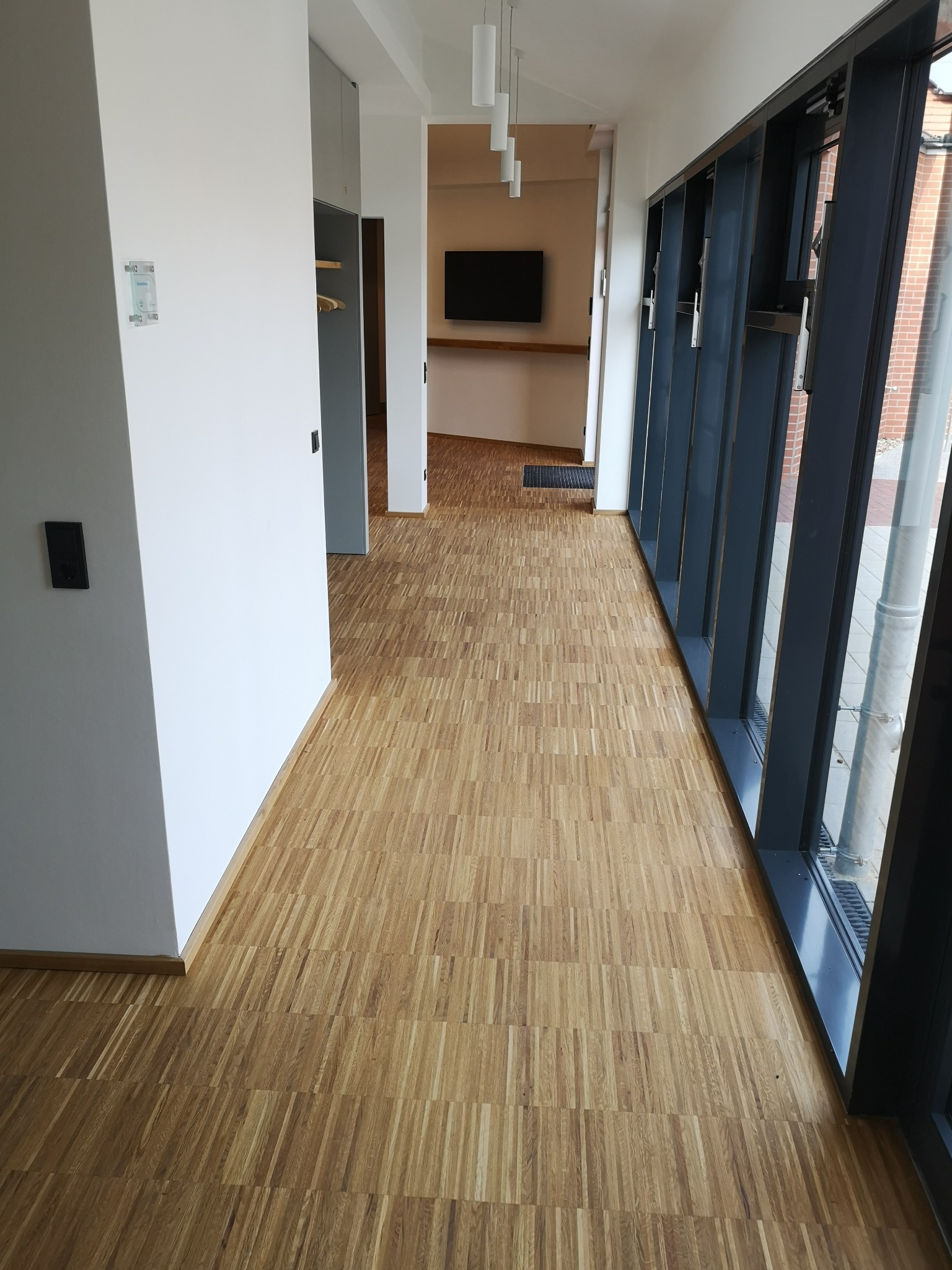 Wnętrze z odnowioną, jasną podłogą w jodełkę, widok z korytarza w Zduńskiej Woli. Duże okna z czarnymi ramami, białe ściany i minimalistyczne oświetlenie.