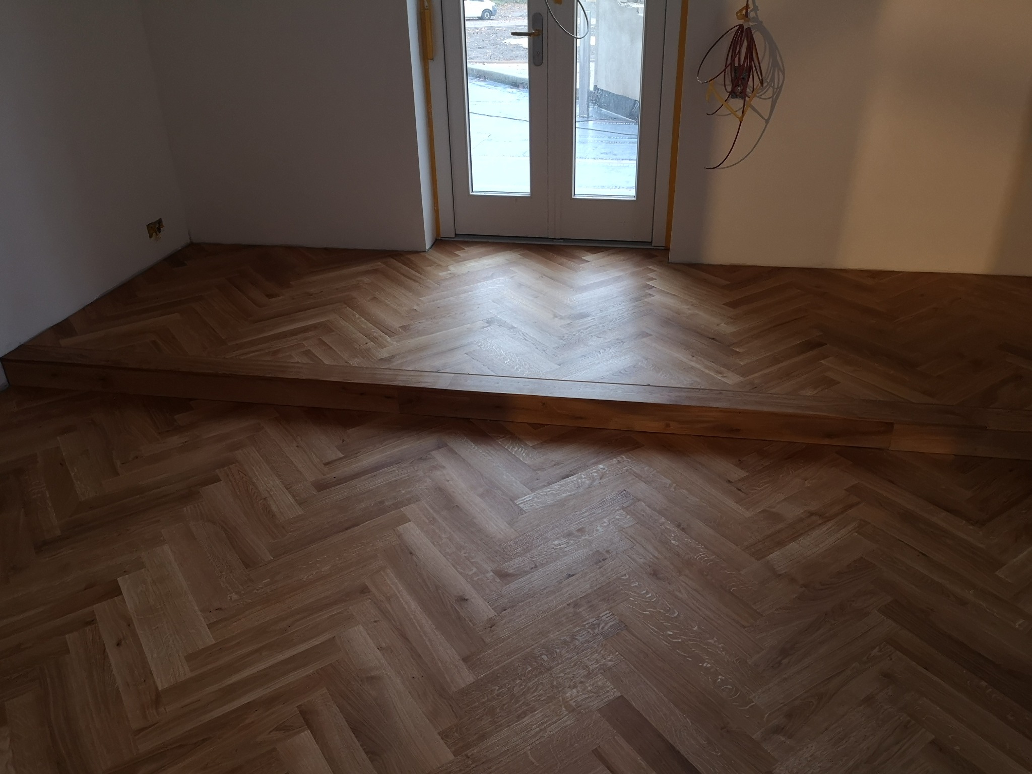 Ułożona podłoga z drewnianego parkietu w jodełkę, widoczny próg i drzwi balkonowe, świeżo po montażu. W tle widoczne kable elektryczne.