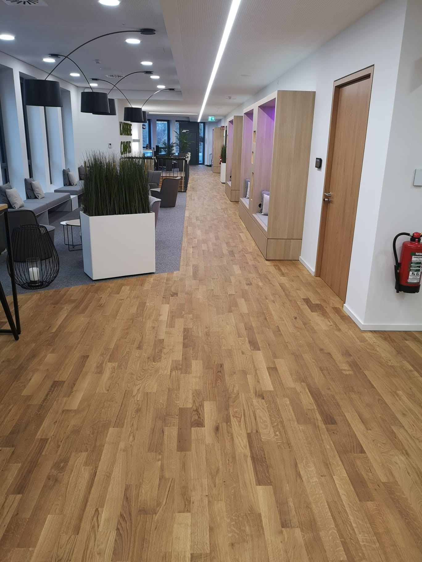 Jasna, drewniana podłoga ułożona w jodełkę w nowoczesnym biurze z elementami zieleni i strefą relaksu. Widoczny fragment gaśnicy na ścianie.