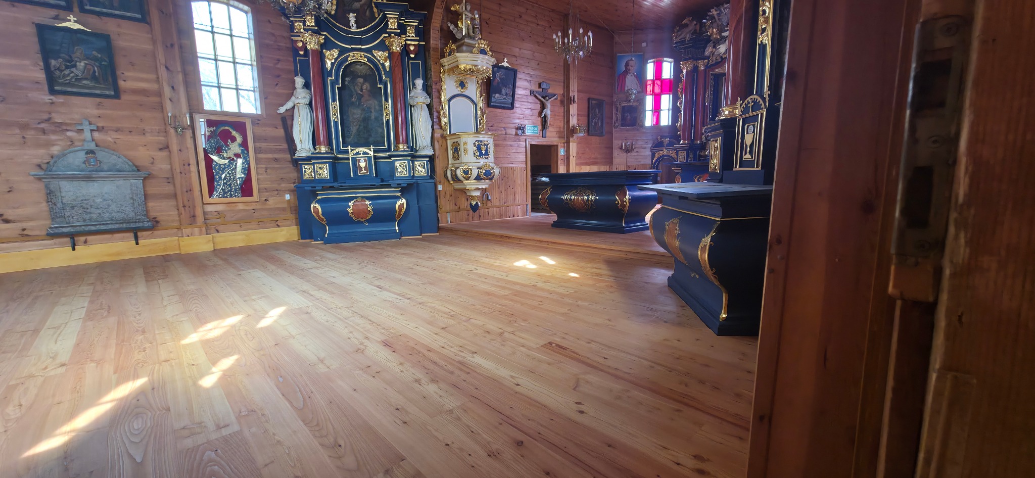 Jasna, drewniana podłoga w zabytkowym kościele, widok zza otwartych drzwi. Na ścianach boazeria, bogato zdobione ołtarze, ambona i obrazy.
