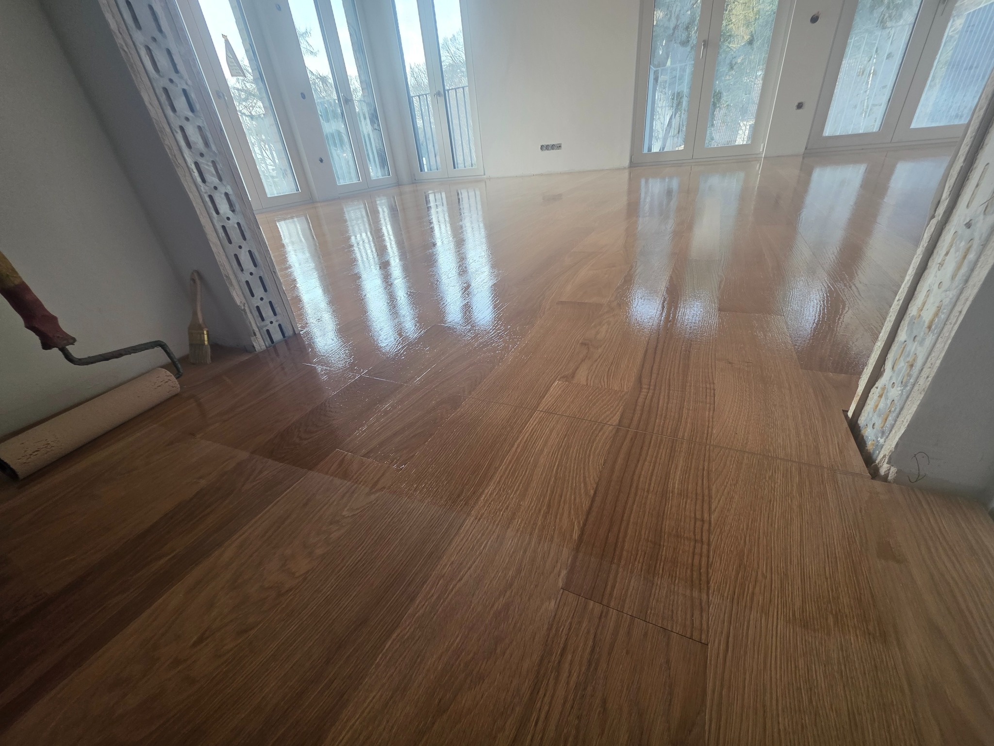 Błyszcząca, świeżo polakierowana drewniana podłoga w jasnym pomieszczeniu z dużymi oknami. Widoczny wałek malarski i pędzel przy ścianie.
