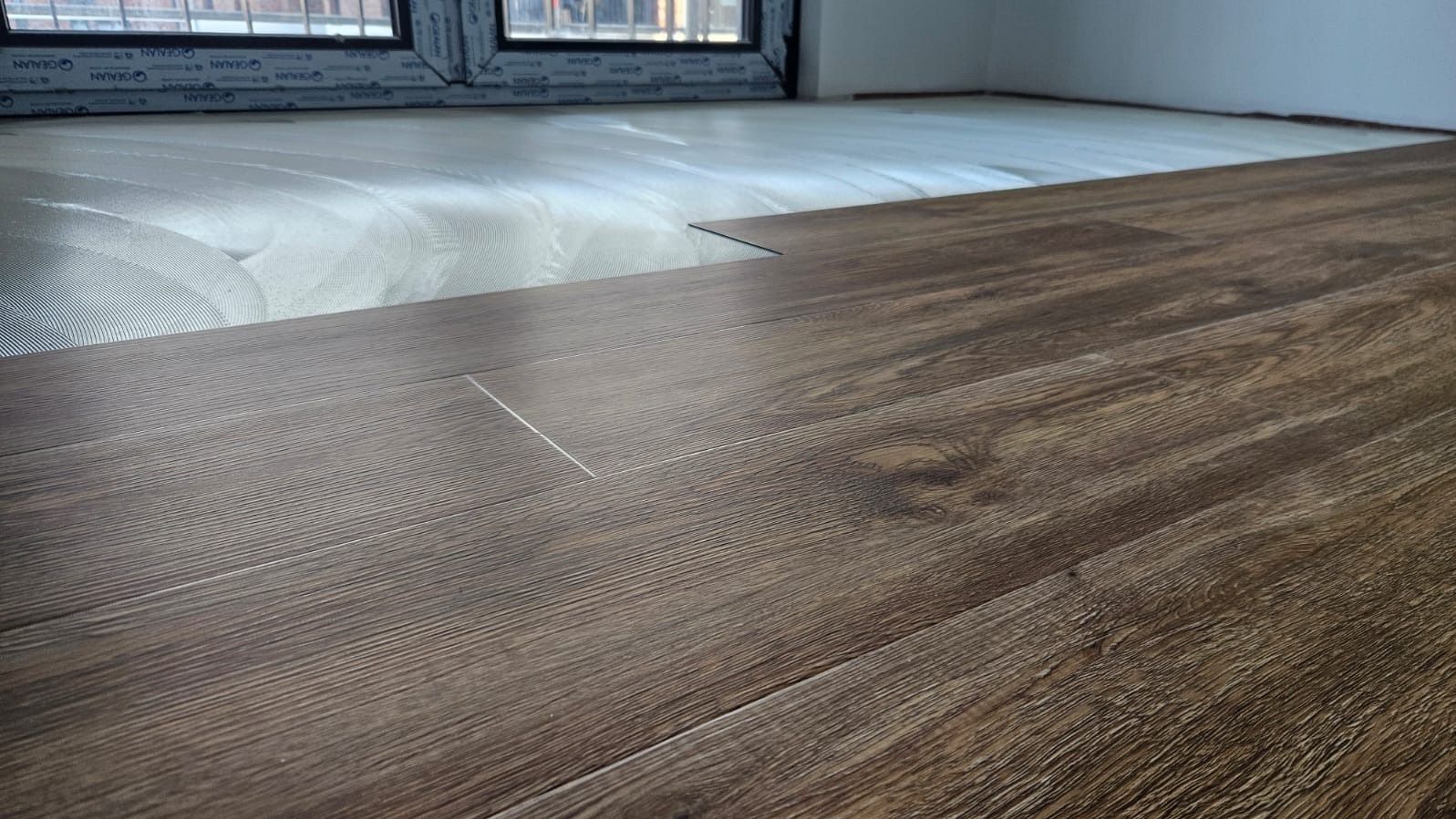 Układanie paneli podłogowych w pomieszczeniu, widoczna warstwa izolacyjna i panele o wzorze drewna, zbliżenie na łączenie paneli przy oknie.
