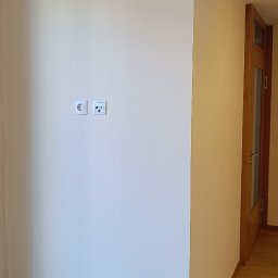 Matpol - Jasna ściana po świeżym malowaniu, z widocznymi gniazdkami elektrycznymi i zabezpieczeniem podłogi. Fragment drewnianej podłogi i drzwi w tle. Neutralne oświetlenie.
