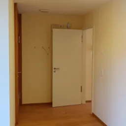 Jasne wnętrze z drewnianą podłogą i białą ścianą. Widoczne drzwi, włączniki i gniazdka elektryczne. Minimalistyczny design, ciepłe oświetlenie sufitowe.